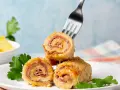 Roladki z piersi kurczaka z szynką i serem, jedna nabita na widelec, na talerzu z natką pietruszki.