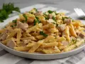 Penne z kremowym sosem i kawałkami indyka, posypane świeżą natką pietruszki. Pyszny makaron z indykiem na talerzu.
