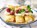 Rolowane naleśniki z mascarpone, udekorowane malinami i miętą, wyglądają apetycznie.