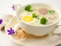 Aromaticzny barszcz biały na żeberkach z jajkiem i kiełbasą, ozdobiony natką pietruszki. Na talerzyku wielkanocne jajka i fioletowe kwiaty.