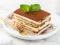 Pyszne tiramisu z mascarpone i śmietaną, posypane kakao i ozdobione miętą. Złoty widelec czeka na pierwszy kęs.