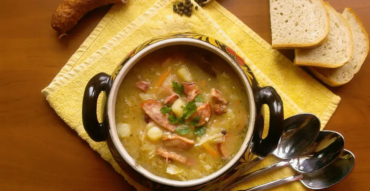 Aromaticzny kapuśniak z kiszonej kapusty, podany w ozdobnym garnku, z kiełbasą, ziemniakami i pieczywem.