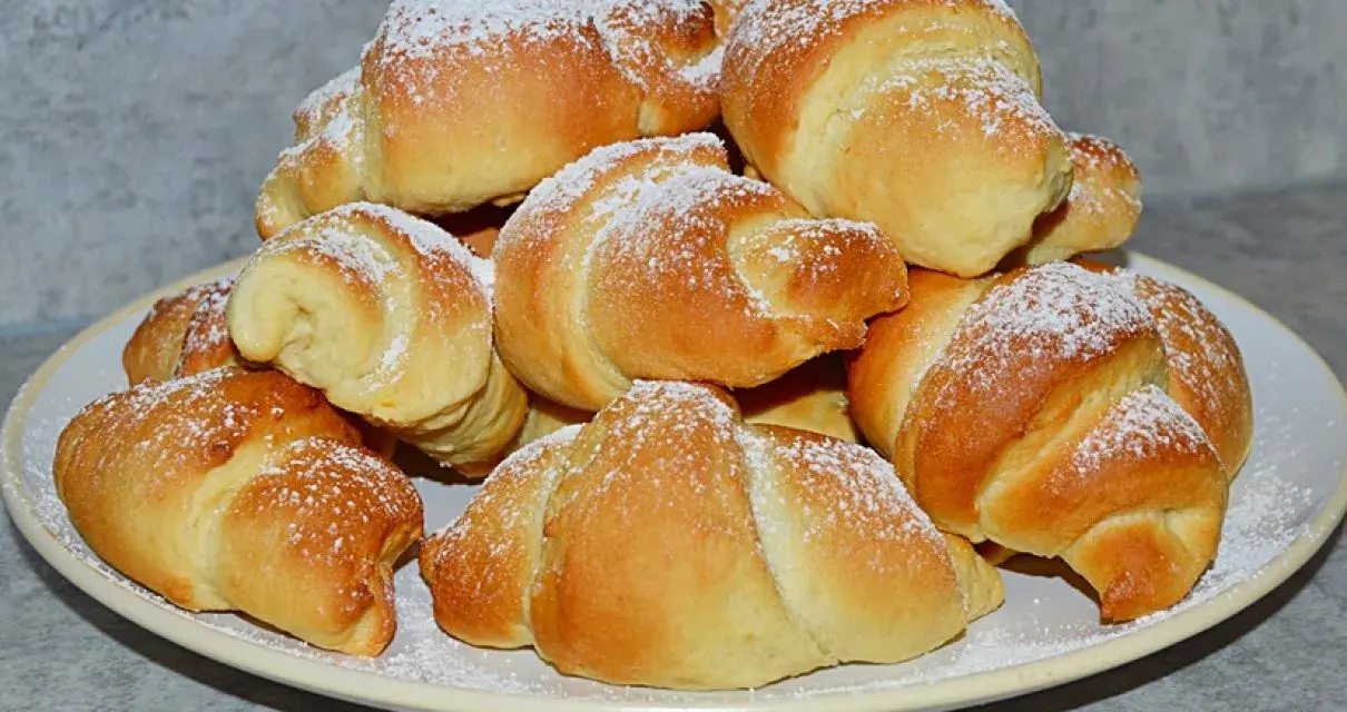 Stos świeżo upieczonych rogalików z czekoladą, posypanych cukrem pudrem, na talerzu.