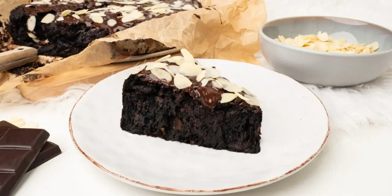 Wilgotny brownie z buraka, polany czekoladą i posypany płatkami migdałów, na talerzu.