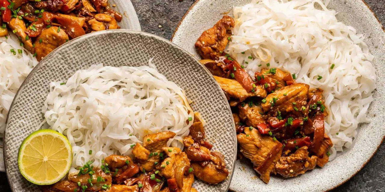 Dwa talerze z kurczakiem teriyaki i makaronem ryżowym, udekorowane plasterkami limonki i szczypiorkiem.