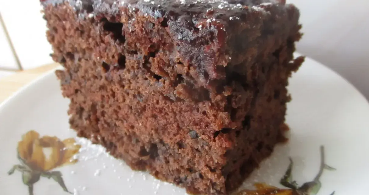 Wilgotne ciasto czekoladowe z dżemem porzeczkowym, posypane cukrem pudrem, na talerzyku z motywem kwiatowym.
