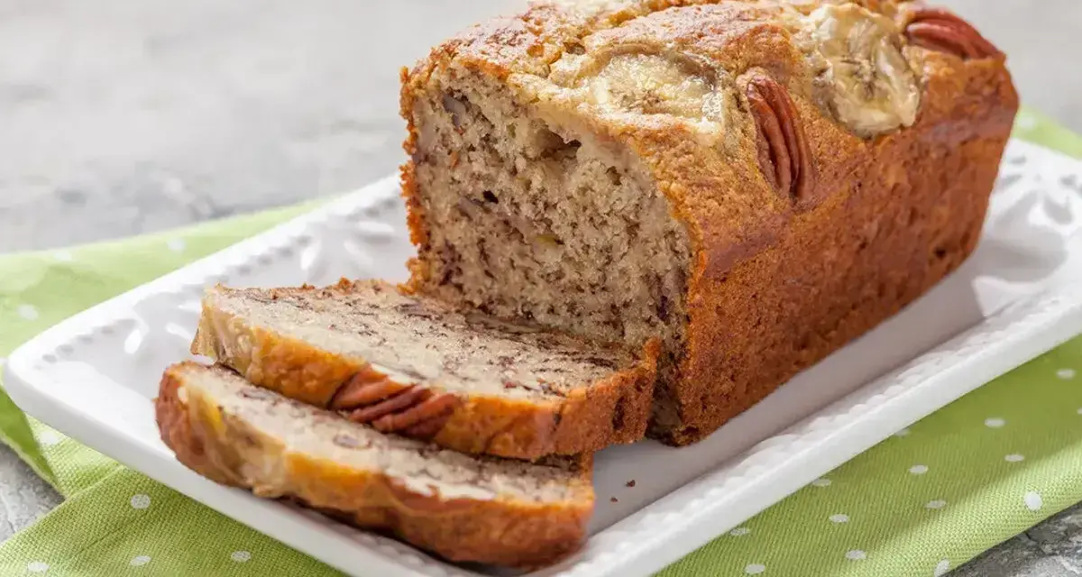 Wilgotny chlebek bananowy z orzechami pekan i plasterkami banana na wierzchu, pokrojony na kromki, podany na białej paterze z zieloną serwetką w groszki.