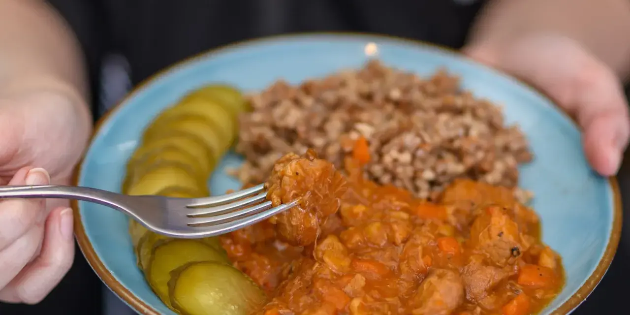 Pyszny gulasz z kaszą gryczaną i ogórkami kiszonymi na talerzu. Widok z bliska na kawałek mięsa nabierany widelcem.