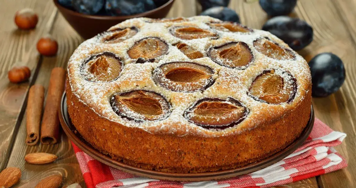 Pyszne ciasto ze śliwkami, posypane cukrem pudrem, na drewnianym blacie. Obok leżą orzechy, migdały i laski cynamonu.
