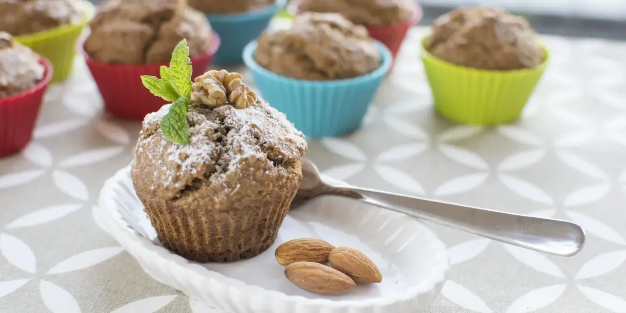 Pyszne muffinki bananowe fit, posypane cukrem pudrem, z orzechami i miętą. Obok migdały i łyżeczka.