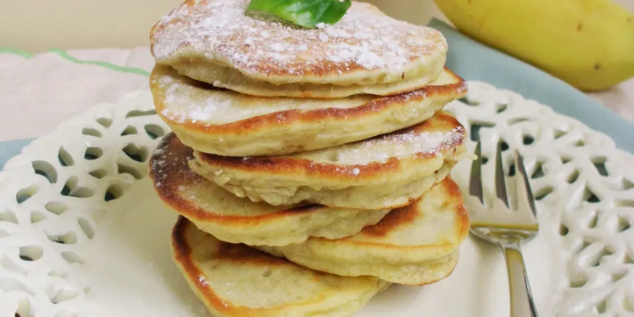 Stosik puszystych placuszków bananowych bez mleka, posypanych cukrem pudrem, z bananem w tle.