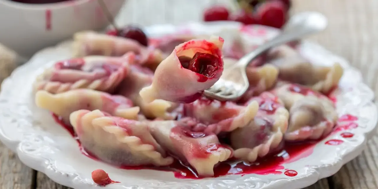 Pyszne pierogi z wiśniami, polane słodkim sosem, kuszą swoim wyglądem. Jedna sztuka jest podniesiona widelcem, ukazując soczyste nadzienie.