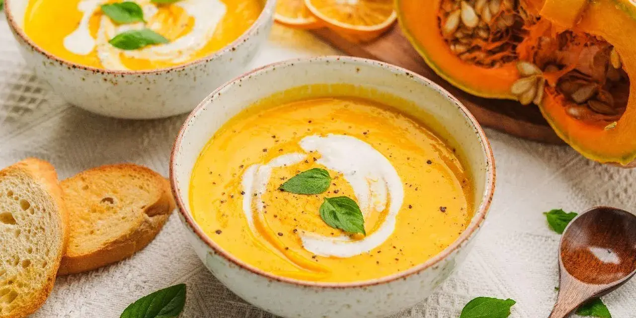 Aksamitna zupa z dyni hokkaido, udekorowana śmietaną i bazylią, podana z grzankami. Obok połówka dyni.