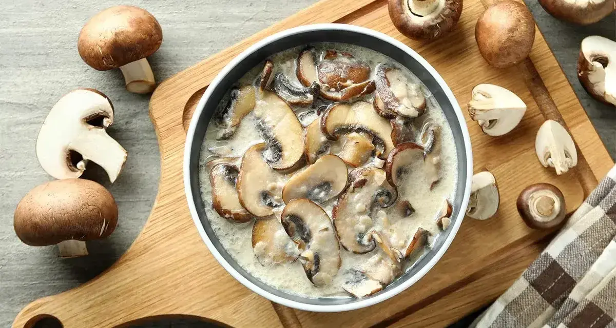 Pyszny sos grzybowy z mrożonych grzybów, podany w misce na drewnianej desce, otoczony świeżymi pieczarkami.