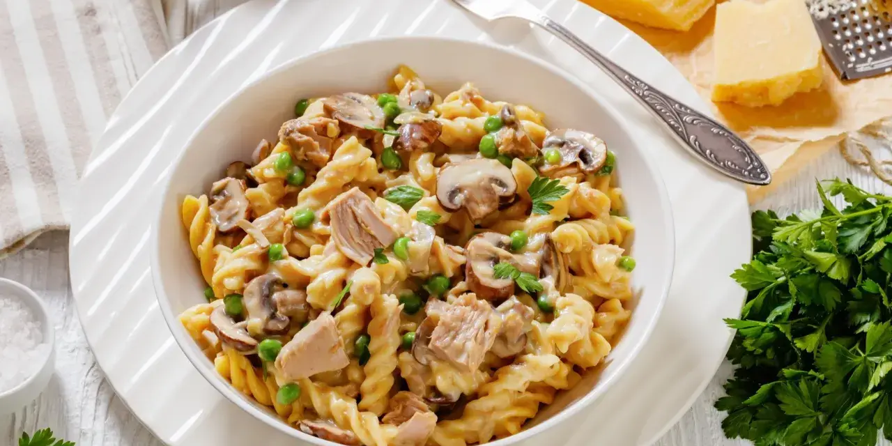 Makaron z tuńczykiem z puszki, groszkiem i pieczarkami w kremowym sosie, posypany natką pietruszki. Obok kawałki parmezanu i tarka.