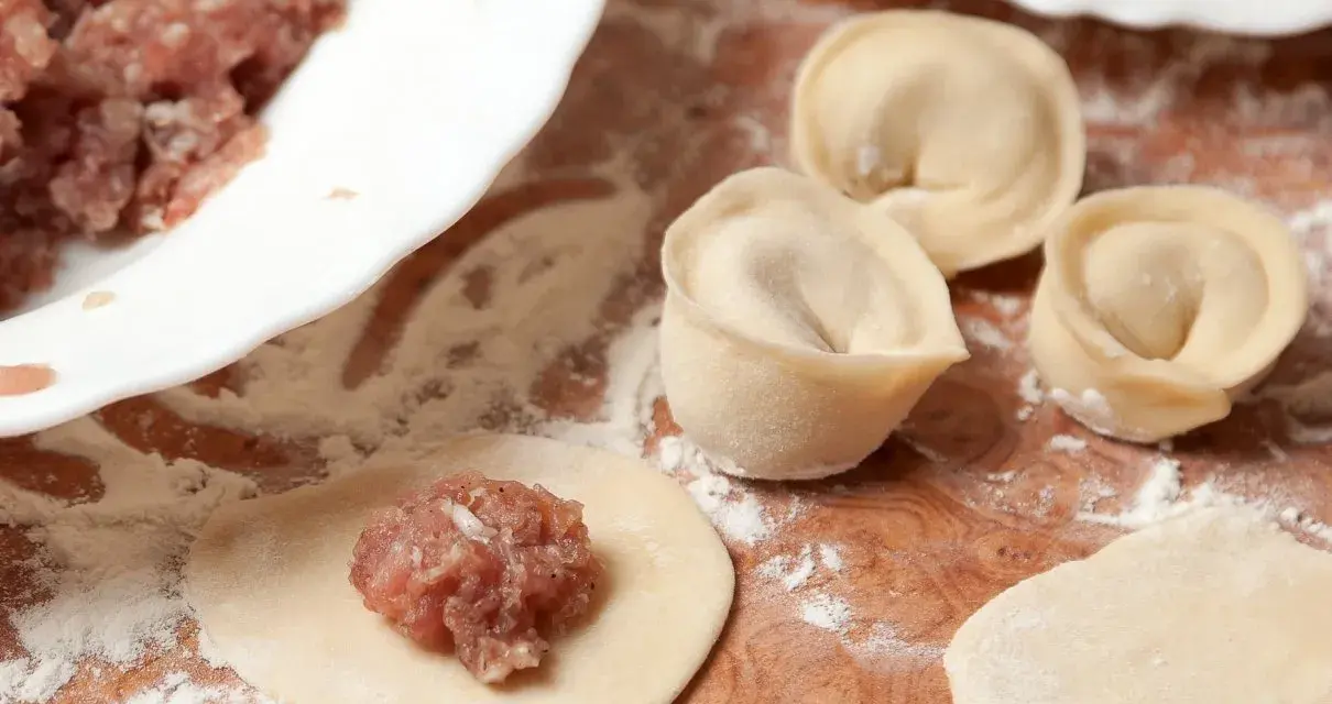 Przygotowanie pierogów z mięsem mielonym surowym. Na drewnianej desce widać surowe ciasto, farsz i gotowe pierożki.