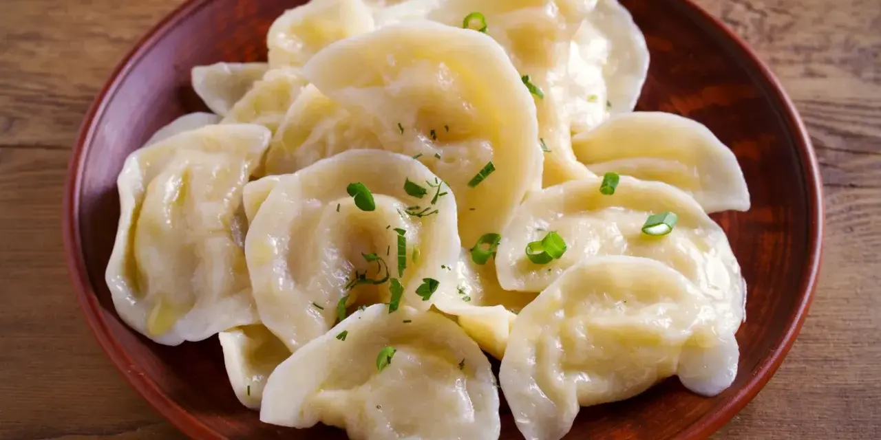 Pyszne pierogi z tradycyjnym farszem do uszek, posypane szczypiorkiem, na glinianym talerzu.