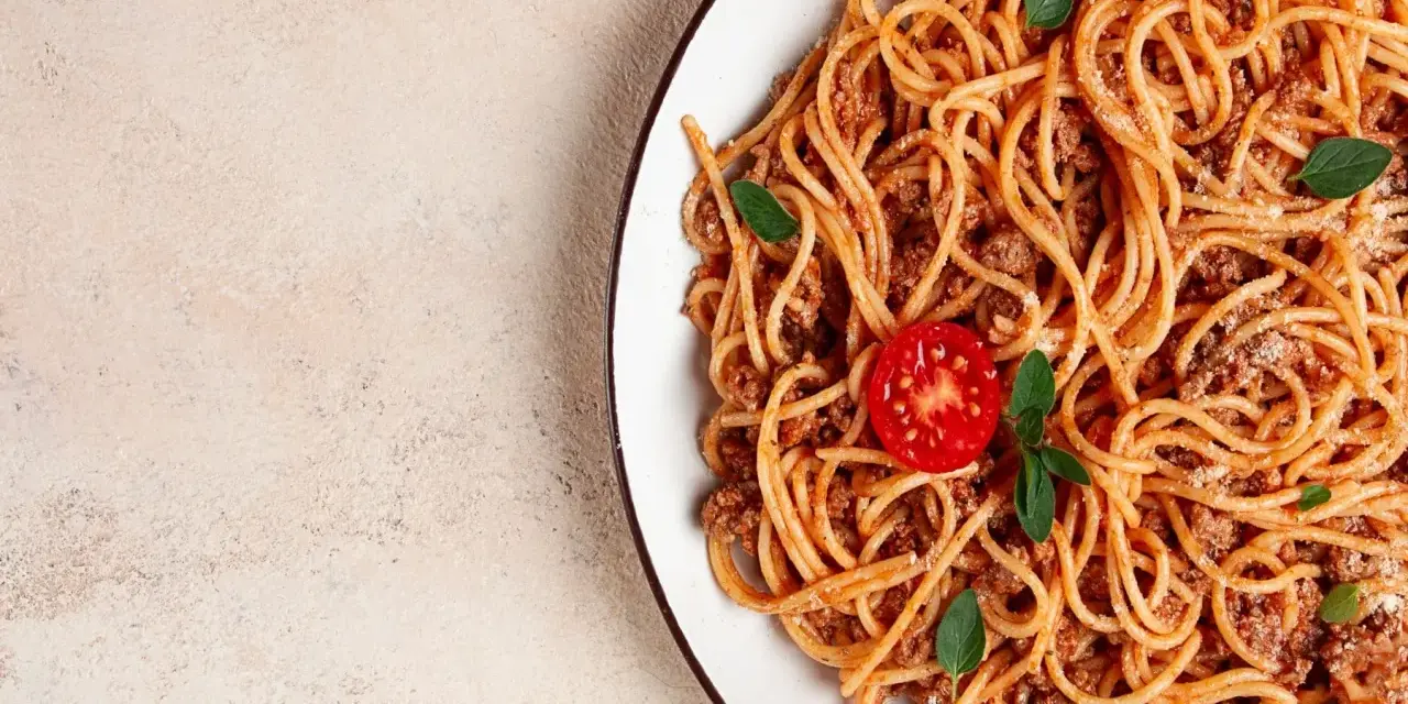 Talerz spaghetti bolognese z pomidorkiem i bazylią na jasnym tle.