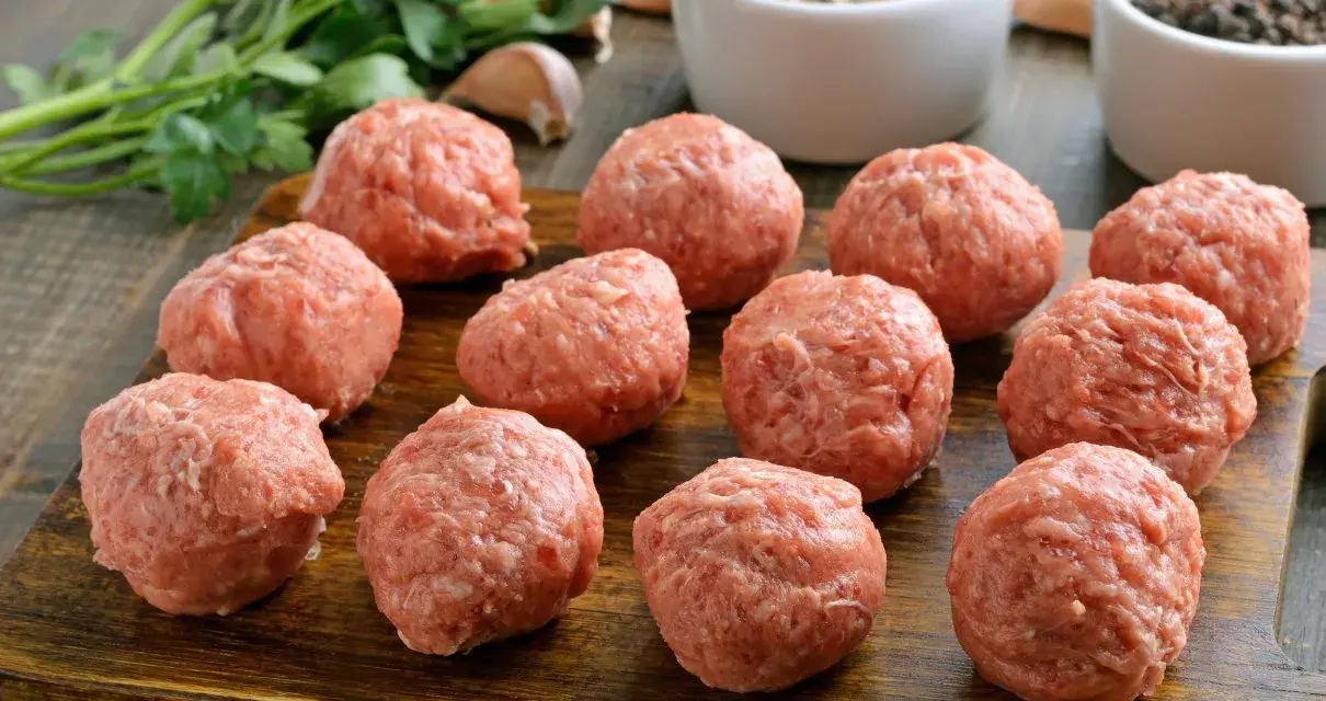 Surowe kotlety mielone uformowane w kulki, gotowe do smażenia. Obok pęczek natki, czosnek i przyprawy.