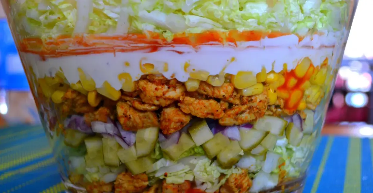 Warstwowa sałatka z kurczakiem w stylu gyros, z kukurydzą, ogórkiem, cebulą i sosem.