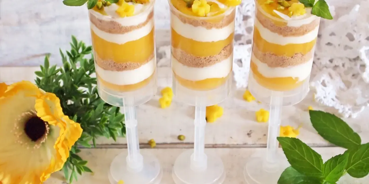 Warstwowy deser z mango w przezroczystych pojemnikach, ozdobiony listkami mięty i pistacjami.