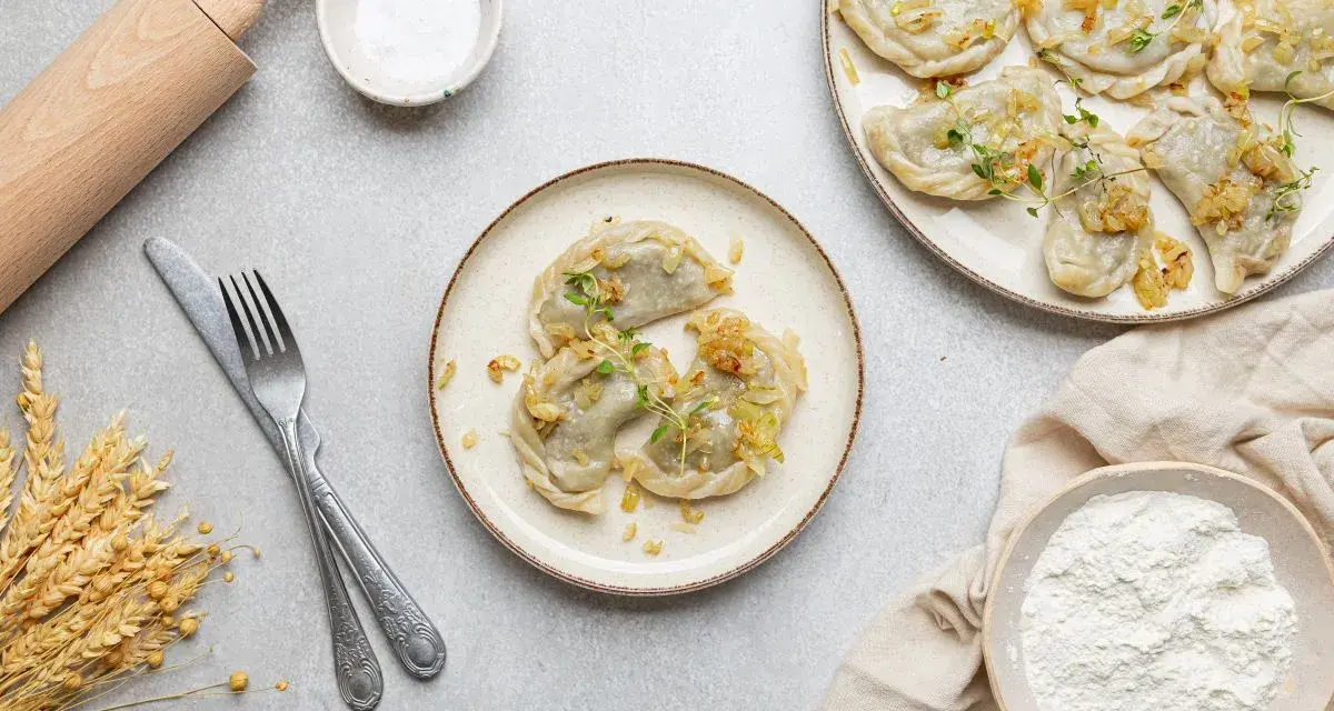 Pyszne pierogi z soczewicą, okraszone cebulką i ziołami, gotowe do podania. Obok wałek, mąka i kłosy zboża.