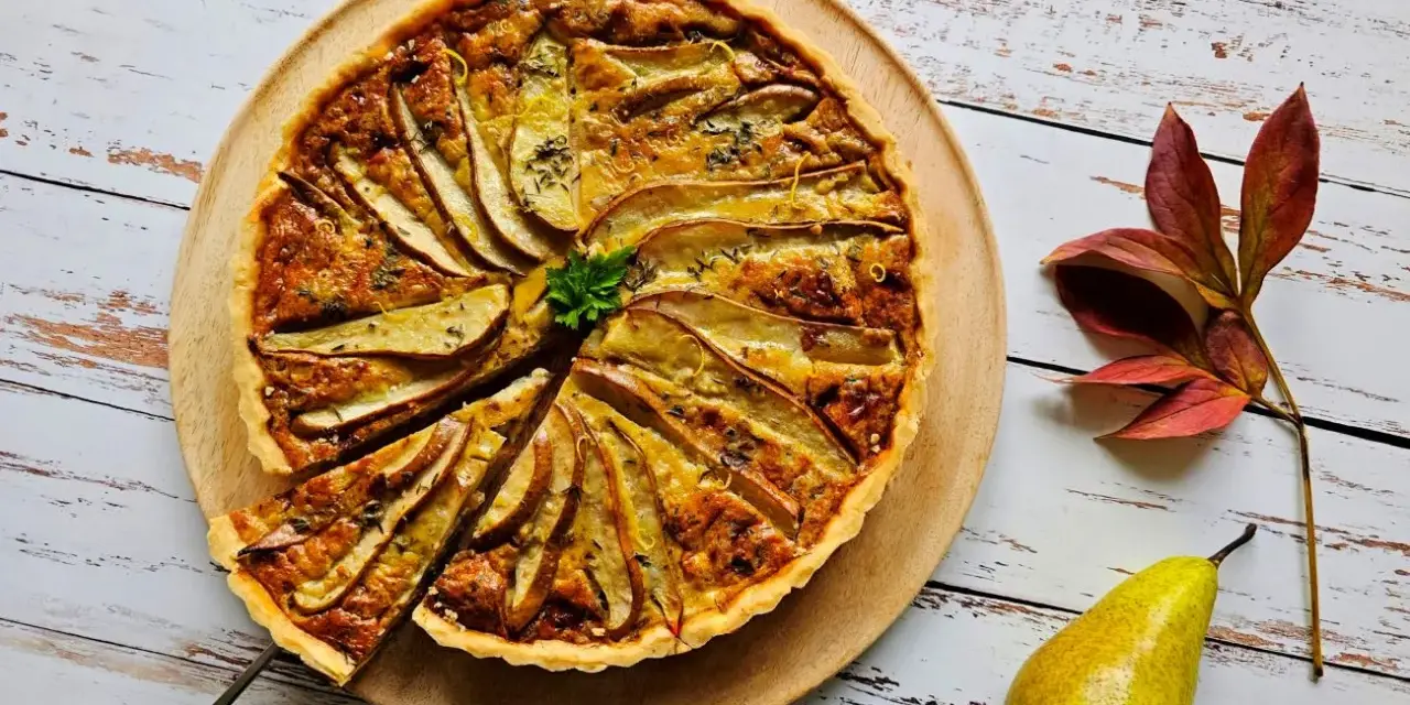 Apetyczna tarta z gruszką i serem pleśniowym, ozdobiona natką pietruszki i skórką cytrynową, na drewnianej desce. Obok leży gruszka i jesienne liście.