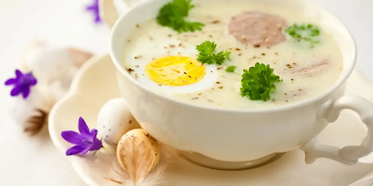 Aromaticzny barszcz biały na żeberkach z jajkiem i kiełbasą, ozdobiony natką pietruszki. Na talerzyku wielkanocne jajka i fioletowe kwiaty.