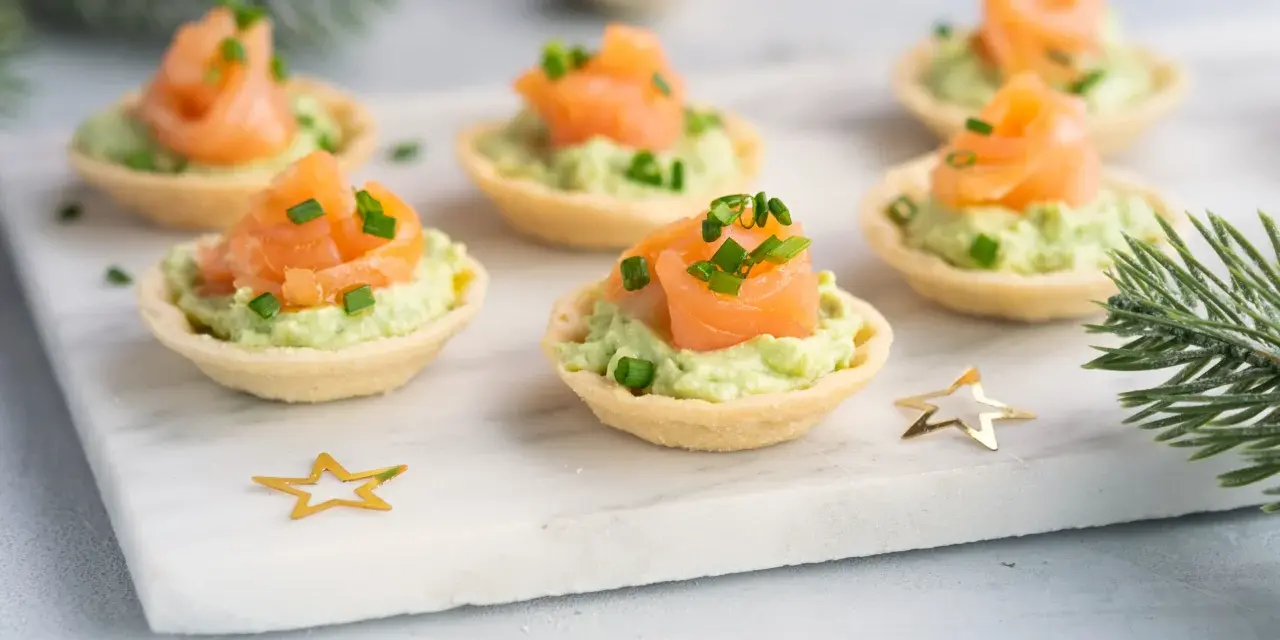 Wytrawne tartaletki z łososiem i awokado, ozdobione szczypiorkiem, na świątecznym tle ze złotymi gwiazdkami i gałązką choinki.