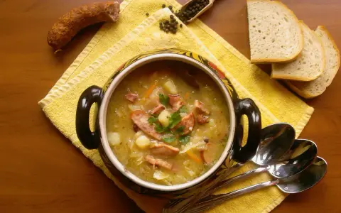 Aromaticzny kapuśniak z kiszonej kapusty, podany w ozdobnym garnku, z kiełbasą, ziemniakami i pieczywem.