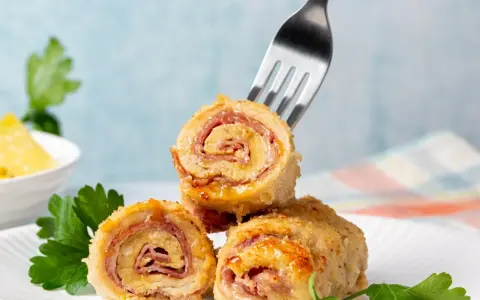 Roladki z piersi kurczaka z szynką i serem, jedna nabita na widelec, na talerzu z natką pietruszki.