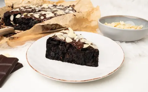 Wilgotny brownie z buraka, polany czekoladą i posypany płatkami migdałów, na talerzu.