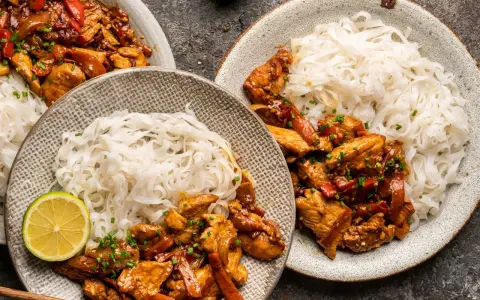 Dwa talerze z kurczakiem teriyaki i makaronem ryżowym, udekorowane plasterkami limonki i szczypiorkiem.