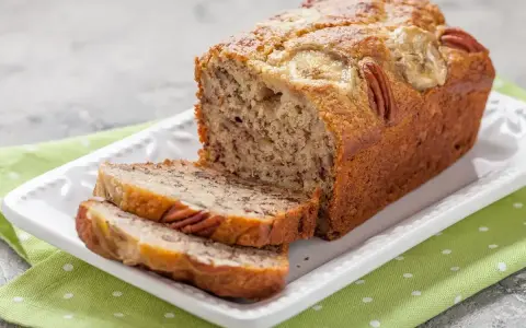 Wilgotny chlebek bananowy z orzechami pekan i plasterkami banana na wierzchu, pokrojony na kromki, podany na białej paterze z zieloną serwetką w groszki.