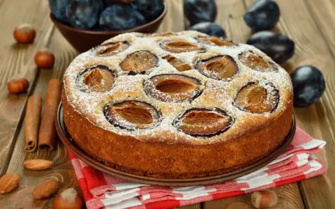 Pyszne ciasto ze śliwkami, posypane cukrem pudrem, na drewnianym blacie. Obok leżą orzechy, migdały i laski cynamonu.