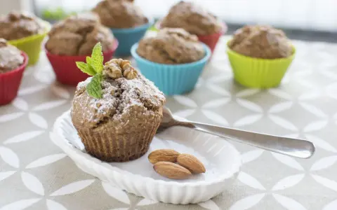 Pyszne muffinki bananowe fit, posypane cukrem pudrem, z orzechami i miętą. Obok migdały i łyżeczka.