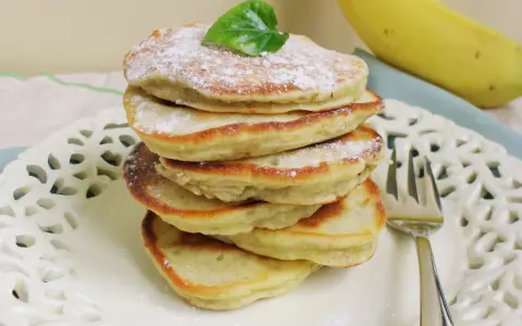 Stosik puszystych placuszków bananowych bez mleka, posypanych cukrem pudrem, z bananem w tle.