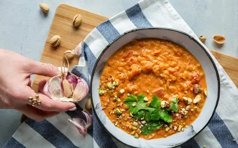 Aksamitna zupa z soczewicy z mlekiem kokosowym, posypana pistacjami i kolendrą. Obok czosnek i orzechy.