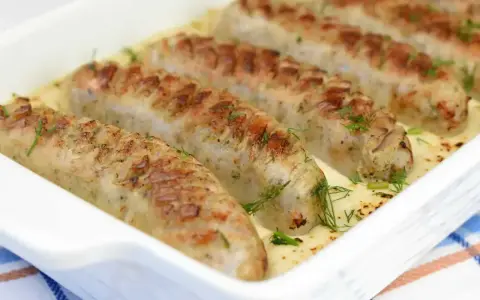 Pieczona biała kiełbasa w kremowym sosie chrzanowym, udekorowana świeżym koperkiem. Pyszne danie gotowe do podania.