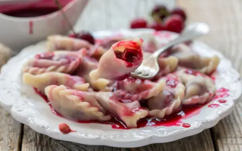 Pyszne pierogi z wiśniami, polane słodkim sosem, kuszą swoim wyglądem. Jedna sztuka jest podniesiona widelcem, ukazując soczyste nadzienie.