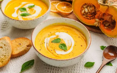 Aksamitna zupa z dyni hokkaido, udekorowana śmietaną i bazylią, podana z grzankami. Obok połówka dyni.