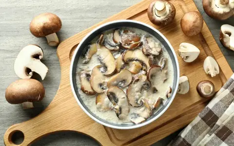 Pyszny sos grzybowy z mrożonych grzybów, podany w misce na drewnianej desce, otoczony świeżymi pieczarkami.