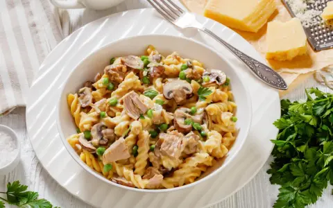 Makaron z tuńczykiem z puszki, groszkiem i pieczarkami w kremowym sosie, posypany natką pietruszki. Obok kawałki parmezanu i tarka.