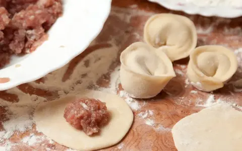 Przygotowanie pierogów z mięsem mielonym surowym. Na drewnianej desce widać surowe ciasto, farsz i gotowe pierożki.
