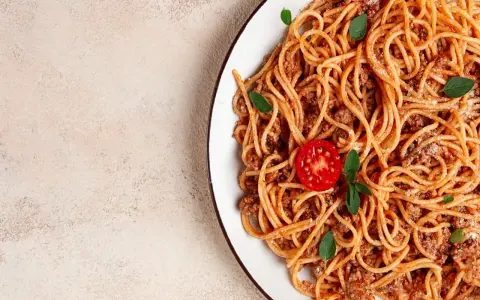 Talerz spaghetti bolognese z pomidorkiem i bazylią na jasnym tle.