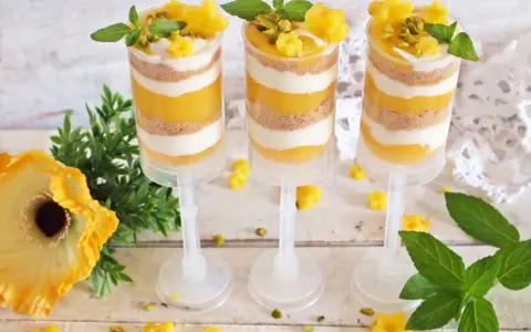 Warstwowy deser z mango w przezroczystych pojemnikach, ozdobiony listkami mięty i pistacjami.