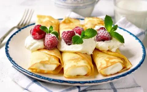 Rolowane naleśniki z mascarpone, udekorowane malinami i miętą, wyglądają apetycznie.