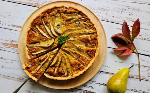 Apetyczna tarta z gruszką i serem pleśniowym, ozdobiona natką pietruszki i skórką cytrynową, na drewnianej desce. Obok leży gruszka i jesienne liście.
