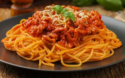 Pyszne spaghetti z mięsem mielonym, polane sosem pomidorowym i posypane startym serem, ozdobione listkiem bazylii.