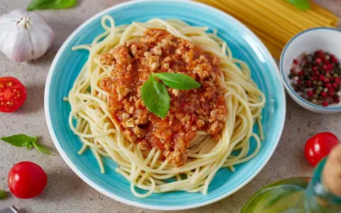 Pyszny, prosty sos do spaghetti z mielonym mięsem i pomidorami, udekorowany bazylią. Obok czosnek, pomidorki i kolorowy pieprz.